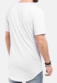 T-shirt en coton blanc avec un ourlet courbé, des manches courtes et un col rond. Marquage subtil au niveau du cou. Porté avec un jean bleu clair.