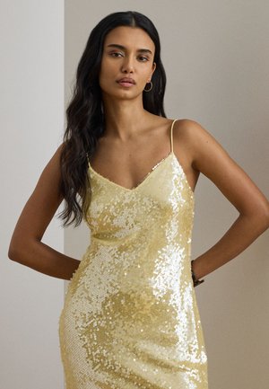 Γυναίκα με μακριά σκούρα μαλλιά, φορώντας χρυσό φόρεμα με παγιέτες και λεπτές τιράντες, στέκεται με τα χέρια στη μέση.