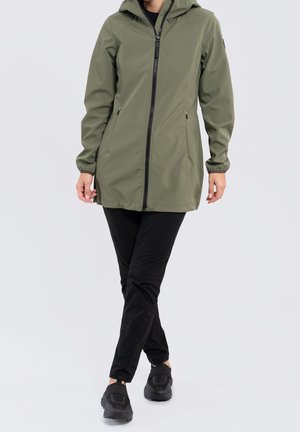 Personne portant une veste imperméable à capuche vert olive fermée, un pantalon noir et des baskets noires, debout devant un fond clair uni.