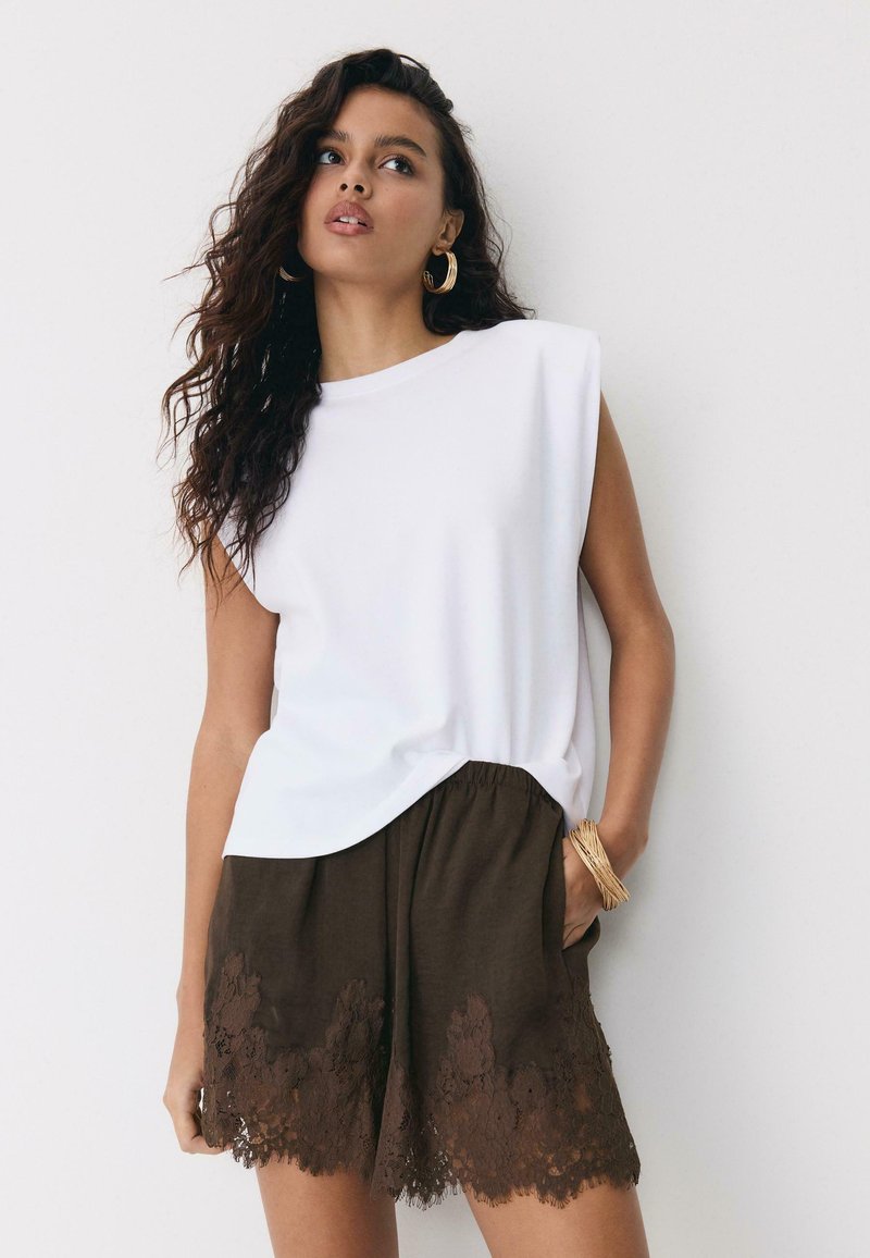 Donna con capelli lunghi e ricci che indossa una canotta bianca senza maniche e pantaloncini marrone scuro rifiniti con pizzo, accessoriati con orecchini a cerchio dorati e bracciale.