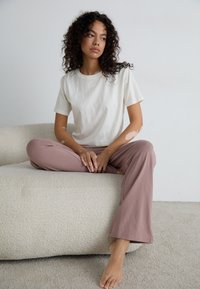 T-shirt beige clair et pantalon rose poussiéreux doux sur une personne assise sur un canapé texturé de couleur crème, pieds nus avec les mains détendues sur les genoux.