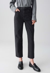 Persona che indossa jeans neri a vita alta e gamba dritta, camicia grigia a maniche lunghe con bottoni e mocassini in pelle nera su uno sfondo neutro.