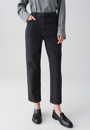 Persoon in zwarte hoge taille rechte jeans, grijze lange mouw knoopshirt, en zwarte leren loafers op een effen achtergrond.