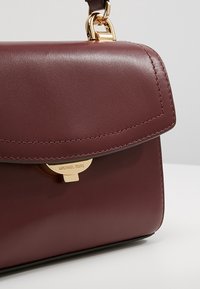 Burgundy læder håndtaske med en guldfarvet lås, der har et struktureret design, glat overflade og kontrasterende syninger langs kanterne.
