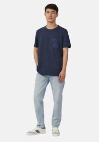 Marineblauwe t-shirt met een zak met een donker bloemenpatroon, gecombineerd met lichtblauwe jeans en witte sneakers met grijze accenten.
