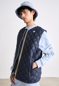 Navy quilted vest with diagonal stitching, white zip, and striped accent. Light blue sweatshirt beneath, worn with a matching bucket hat.