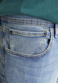 Blue denim jeans with a classic five-pocket design. Features include a back pocket with a metal button and visible stitching details.