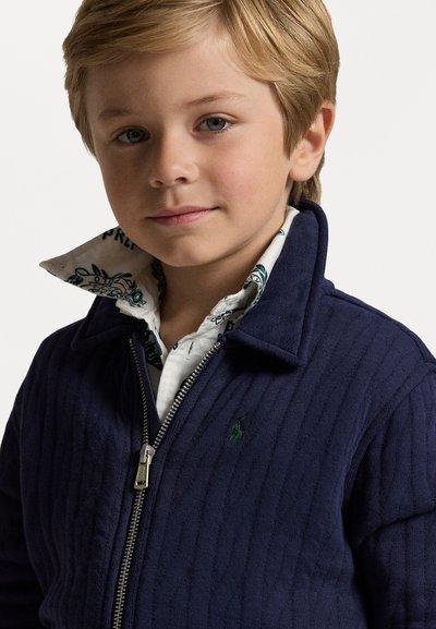 Casaco azul marinho com nervuras, fecho de correr e gola alta, com um logotipo verde. Sobrepõe-se a uma camisa branca estampada. Textura suave, design casual.