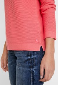 Koraliowy różowy sweter z prążkowanej tkaniny, z rękawami o długości trzech czwarci, z bocznym rozcięciem i małym różowym guzikiem na dole, zestawiony z niebieskimi dżinsami.