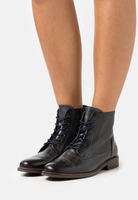 Bottes noires en cuir à la cheville avec un design nez de cap, comportant des lacets bleus, un col légèrement plus haut et une semelle plate marron.