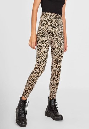 Femme portant un pantalon taille haute à imprimé léopard, un haut noir sans manches et des bottes de combat noires lacées, debout avec une jambe légèrement pliée.