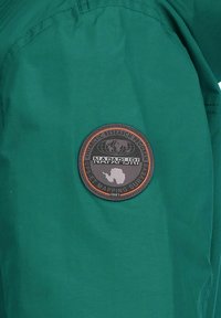 Grønt stof med en glat tekstur har et rundt sort og orange Napapijri-logo med et motiv af en globus og Antarktis.
