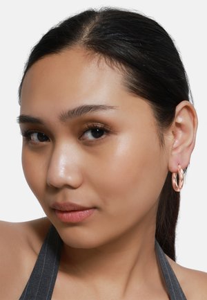 Roségoldene Creolen mit einem gedrehten Design, prominent an einem Modell mit dunklem Haar, das ein grau gestreiftes Oberteil trägt.
