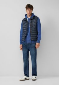 Giovane uomo che indossa un felpa blu con cappuccio, un gilet trapuntato nero, jeans blu e sneakers bianche, in piedi di fronte a uno sfondo neutro semplice.