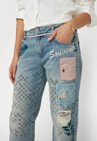 Šviesiai mėlynos džinsinės kelnės su įvairiais priedais, įskaitant rožinį flamingą, gėlių raštus ir siuvinėjimą "Suvenyras", su nusidėvėjimo detalėmis.