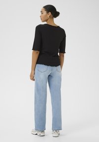 Sort ribbet top med bølget kant og ærmer, parret med lyseblå brede jeans. Model iført hvide sneakers, set fra bagenden.