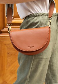 Hnedá kožená crossbody taška s okrúhlym tvarom, plochým vrchom, čierno-bielym pruhovaným popruhom a embosovaným logom na prednej strane.