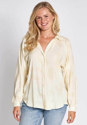 Cream-colored, long-sleeve button-up shirt with a soft texture and abstract light patterns; paired with light blue jeans.
