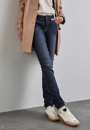 Jean bleu foncé à coupe slim, accompagnés de baskets blanches et beiges. Un trench-coat beige superposé à un pull léger vient parfaire l'ensemble.
