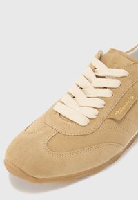 Sneaker in suede di un beige chiaro con accenti testurizzati beige e lacci color crema. Presenta una punta arrotondata e una suola piatta in gomma color tan.