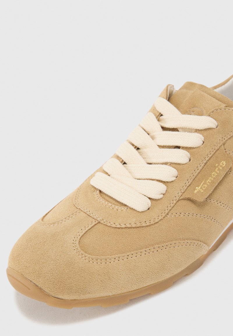 Skórzane sneakersy w jasnym beżu z beżowymi teksturowanymi akcentami i kremowymi sznurówkami. Mają okrągły czubek i płaską, brązową gumową podeszwę.