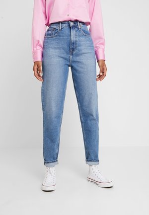 Person trägt hellblaue, hoch taillierte Jeans, die an den Knöcheln hochgekrempelt sind, ein pinkes Langarmshirt und weiße Schnürschuhe.