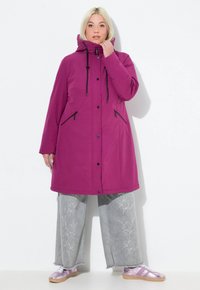 Woman wearing a long purple hooded coat, light gray wide-leg jeans with floral embroidery, and metallic pink sneakers standing against a white wall.