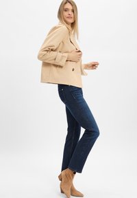 Beige doppelt geknöpfter Blazer mit großen Knöpfen, dunkelblaue gerade Jeans und hellbraune Wildleder-Ankle Boots, auf einem neutralen Hintergrund posiert.