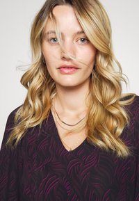 Tecido escuro com um padrão roxo em espiral, apresentando um design em V. Três colares de ouro em camadas adornam o decote. Cabelo suave e ondulado.