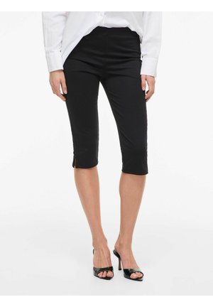 Femme portant un pantalon noir slim longueur genou avec petites fentes latérales, une chemise blanche à manches longues et des escarpins noirs à bout ouvert.