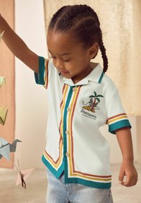 Short-sleeved white shirt with green, orange, and yellow stripes, featuring a palm tree graphic and brown buttons. Worn with blue denim shorts.