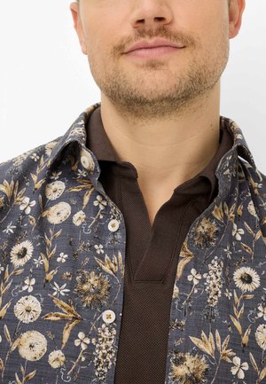 Mann mit leichtem Gesichtshaar, der ein dunkelbraunes Poloshirt unter einem blau geblümten Hemd vor weißem Hintergrund trägt.