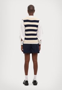 Kootud vest tumesinise ja kreemja horisontaalse triibuga, millel on ribikant ja õlapaneelid. Kombineeritud tumesiniste lühikestega ja mustade kingadega.