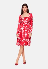 Rotes Kleid mit langen Ärmeln, quadratischem Ausschnitt und weißen Blumenmuster. Glatter Stoff, schwingender Rock und ergänzt durch rote Absatzschuhe.