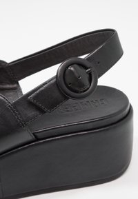 Černé kožené platformové sandály s širokým nártem, který má kruhovou černou přezku a texturovanou stélku. Zvýšená platforma.