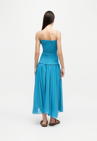 Femme aux longs cheveux foncés portant un haut smocké bleu et une jupe bleue fluide, debout pieds nus sur un fond blanc, tournée de dos.