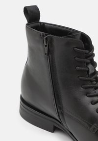 Bottine en cuir noir à bout rond, à lacets sur le devant et avec une fermeture éclair sur le côté. Présente un dessus texturé et un talon empilé.