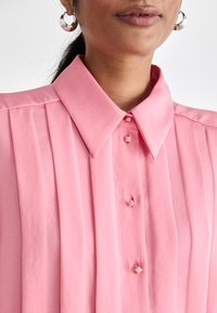 Rosa plissert bluse med spiss krage, prydet med firkantede knapper og en glatt tekstur. Sølvfargede sirkulære øredobber kompletterer utseendet.
