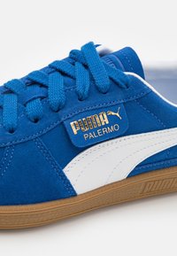 Baskets Puma Palermo bleues avec bande blanche, logo Puma doré, lacets bleus et semelle en gomme sur fond blanc.