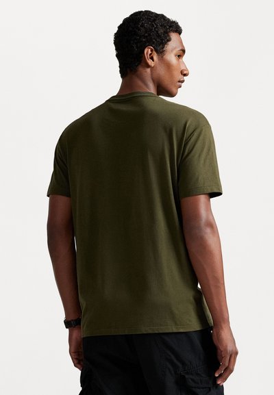 Bărbat cu păr creț scurt, îmbrăcat cu un tricou verde măsliniu și pantaloni cargo negri, întors ușor pe o parte, pe un fundal simplu.