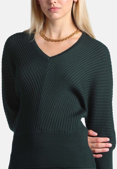 Femme portant un pull en maille côtelée vert foncé et un gros collier chaîne en or, une main posée sur sa taille.