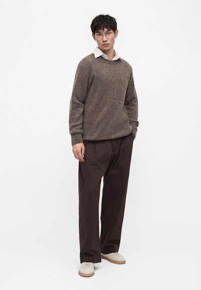 Jeune homme portant un pull marron clair par-dessus une chemise blanche, pantalon large marron foncé, baskets blanches, debout sur un fond blanc uni.
