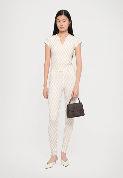 Twee-delige outfit met een geprint top en leggings in crème met een bruin geometrisch ontwerp. Vergezeld van een kleine bruine handtas en witte hakken.