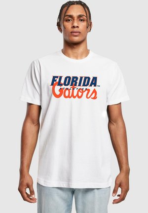 T-shirt blanc en coton avec un texte imprimé en bleu et orange audacieux portant "FLORIDA GATORS" sur la poitrine. Coupe décontractée avec manches courtes.