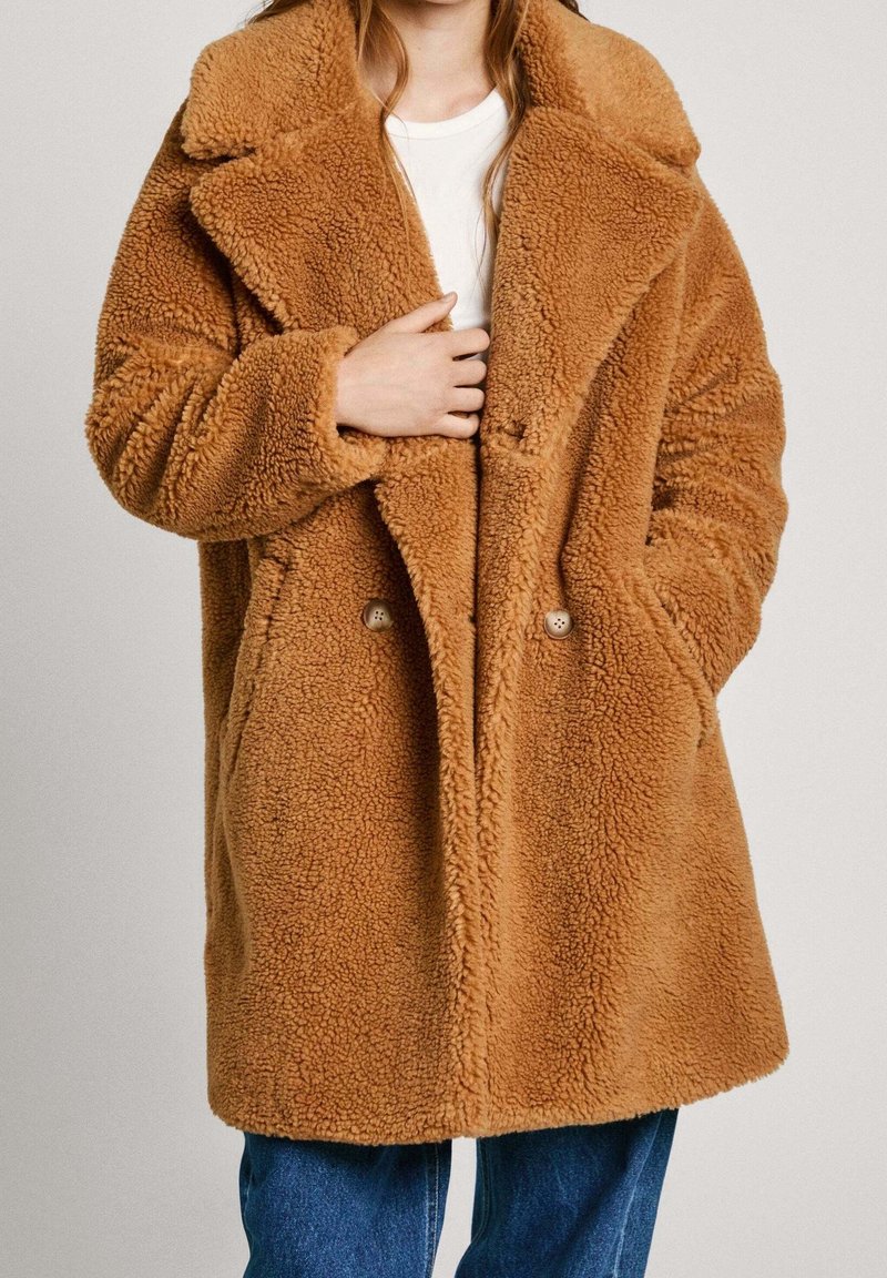 Brązowy, puszysty płaszcz z wełny z wyciętym kołnierzem i dwiema guzikami. Posiada luźny krój i boczne kieszenie, zestawiony z dżinsami.