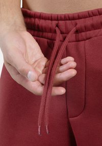 Vinrøde joggingbukser med elastisk taljebånd, udstyret med flettede snore og sidelommer. Glat stof med en let tekstureret overflade.