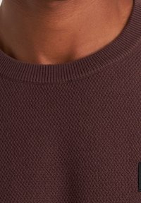Pull marron texturé avec un col côtelé et un patch logo noir, présentant un motif en tricot tissé et une finition de tissu lisse.