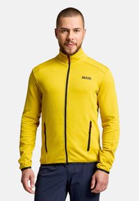 Man with short hair and beard wearing a bright yellow zip-up jacket and dark pants, standing against a light background.