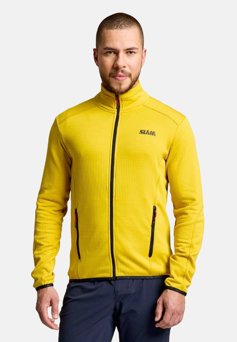 Man with short hair and beard wearing a bright yellow zip-up jacket and dark pants, standing against a light background.