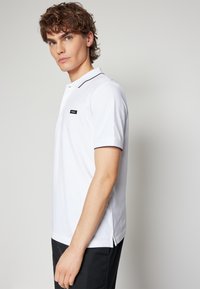 Polo bianco con bordi neri, maniche corte, fenditure laterali all'orlo, con un piccolo etichetta logo nera sul petto.